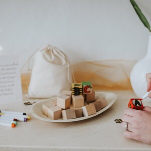 May include: A collection of wooden alphabet blocks, some with painted letters, arranged on a white oval dish. A person is using a red marker to write on a small red wooden block. A white drawstring bag and a card with text are also present.