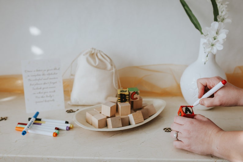 May include: A collection of wooden alphabet blocks, some with painted letters, arranged on a white oval dish. A person is using a red marker to write on a small red wooden block. A white drawstring bag and a card with text are also present.