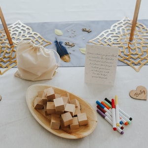 May include: A baby shower setup with wooden blocks, a wooden heart that says "Baby", and a card with instructions. A small drawstring bag and a wooden tray with blocks are also present. Colorful markers are arranged nearby.