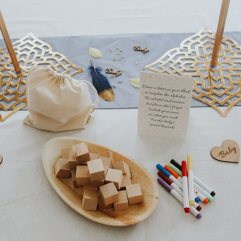 May include: A baby shower setup with wooden blocks, a wooden heart that says "Baby", and a card with instructions. A small drawstring bag and a wooden tray with blocks are also present. Colorful markers are arranged nearby.