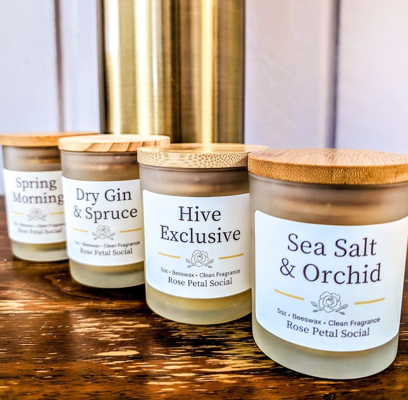 May include: Four glass candle jars with wooden lids. The candles are labeled "Spring Morning", "Dry Gin & Spruce", "Hive Exclusive", and "Sea Salt & Orchid". Each jar has a label with the text "Rose Petal Social" and "5oz Beeswax Clean Fragrance".