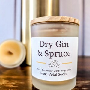 May include: A frosted glass candle jar with a wooden lid. The candle is labeled "Dry Gin & Spruce" and "5oz. Beeswax. Clean Fragrance. Rose Petal Social."