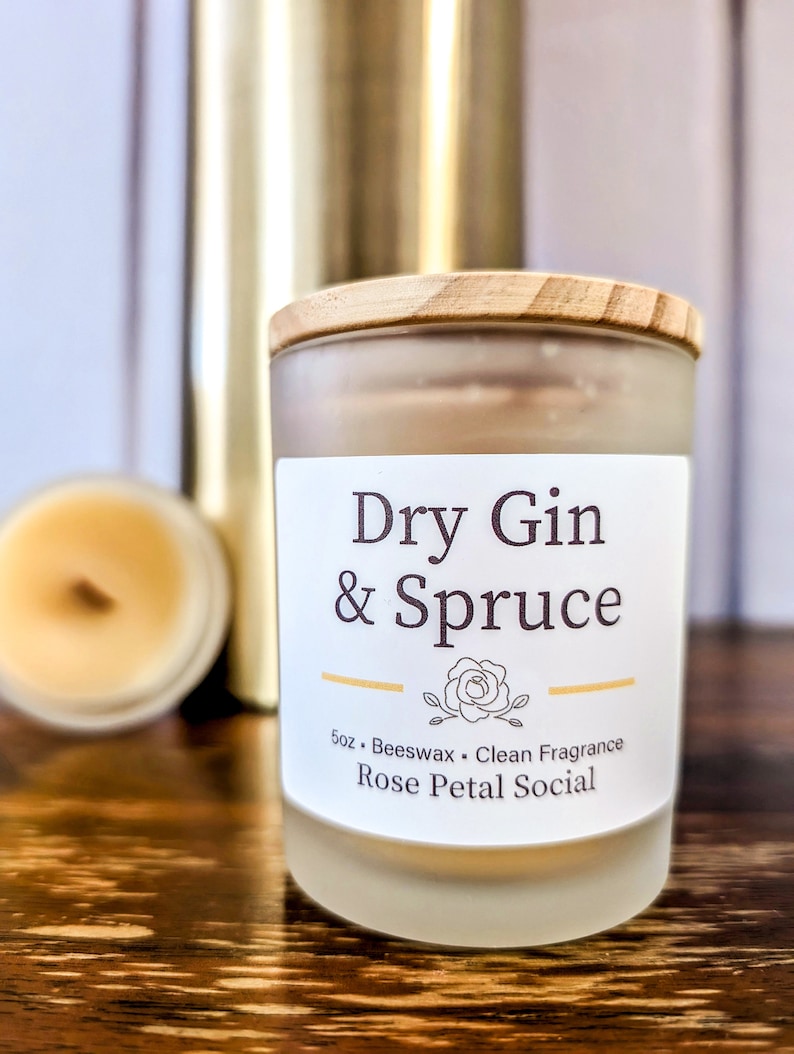 May include: A frosted glass candle jar with a wooden lid. The candle is labeled "Dry Gin & Spruce" and "5oz. Beeswax. Clean Fragrance. Rose Petal Social."