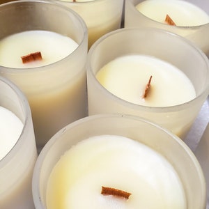 May include: A group of six clear glass candle jars with white wax and wooden wicks. The candles are arranged in a semi-circle.