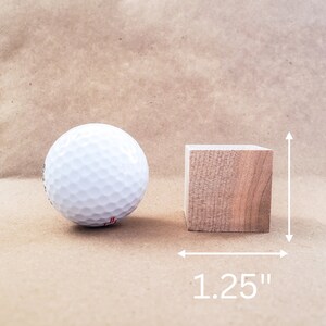 May include: A white golf ball next to a small, natural wood cube. The cube measures 1.25 inches on each side. The background is a solid, light brown color.