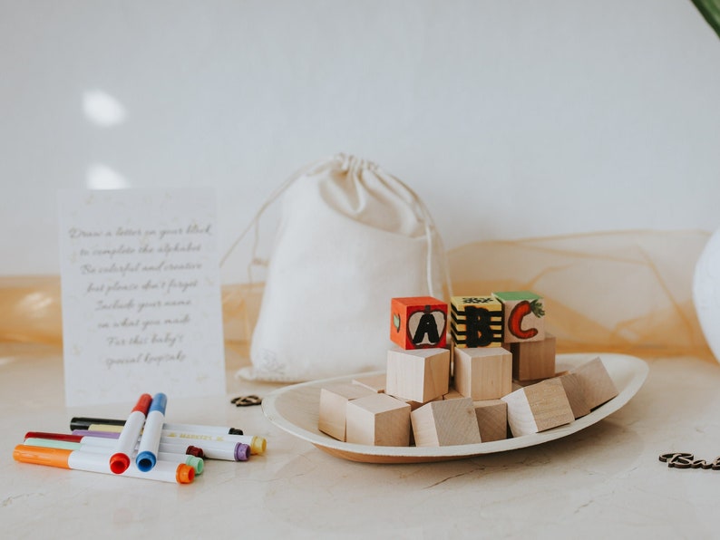 May include: Wooden alphabet blocks with the letters A, B, and C, displayed on a wooden tray. A small drawstring bag and a card with handwritten text are also present. Colorful markers are arranged in the foreground.