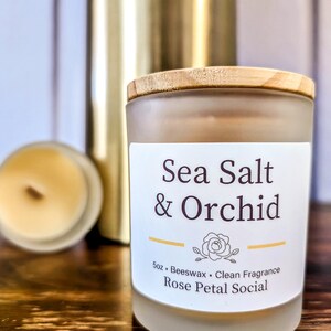 May include: A frosted glass candle jar with a wooden lid. The candle is labeled "Sea Salt & Orchid" and "Rose Petal Social". The candle is 5 oz and made with beeswax.