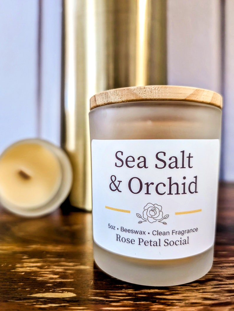 May include: A frosted glass candle jar with a wooden lid. The candle is labeled "Sea Salt & Orchid" and "Rose Petal Social". The candle is 5 oz and made with beeswax.