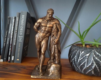 Puede incluir: Una estatua de bronce de una figura masculina desnuda de pie sobre una roca. La figura tiene barba y sostiene un trozo de tela en su mano izquierda. La estatua está en un estante de madera con libros detrás.