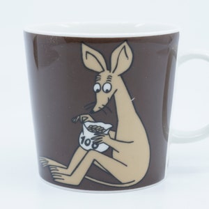 Puede incluir: Una taza de cerámica marrón con interior y asa blancos. La taza presenta una ilustración de dibujos animados de una criatura marrón claro sosteniendo una bolsa de frijoles. La bolsa es blanca con el número 10.
