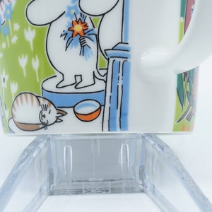 May include: White ceramic mug with a wraparound illustration featuring a cartoon character holding flowers, a sleeping cat, and a bowl. The background is green with white flowers. The mug has a white handle.