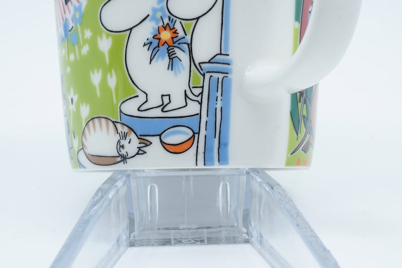 May include: White ceramic mug with a wraparound illustration featuring a cartoon character holding flowers, a sleeping cat, and a bowl. The background is green with white flowers. The mug has a white handle.