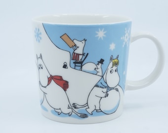 May include: A white ceramic mug with a blue background and a winter scene featuring Moomins making snowmen. The mug has a white handle.