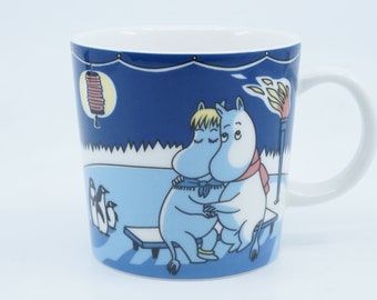 May include: A blue ceramic mug with a white handle features a winter scene with two cartoon characters, a red lantern, and penguins. The characters are hugging and wearing scarves. The text "Moomin" is visible on the mug.