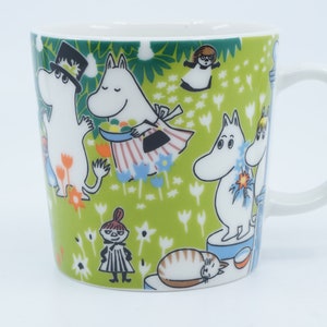 May include: White ceramic mug with a green background featuring cartoon characters. The mug has a white handle and is decorated with various characters and floral patterns. The design includes a cat, and other characters.