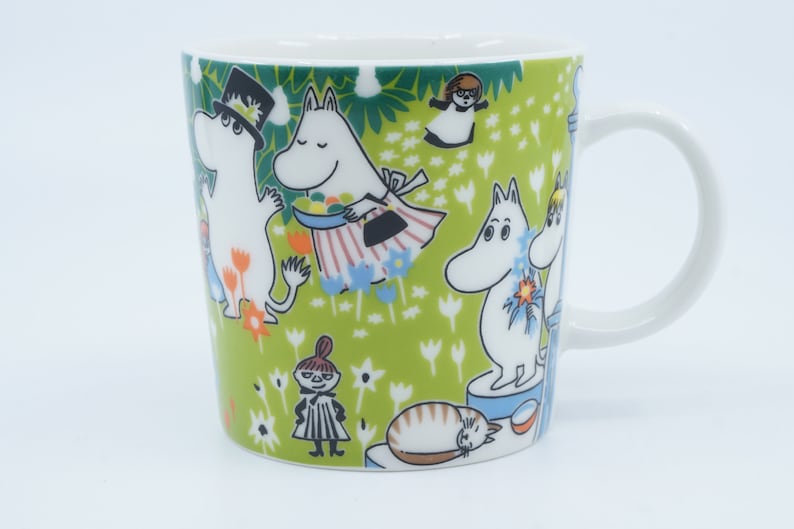 May include: White ceramic mug with a green background featuring cartoon characters. The mug has a white handle and is decorated with various characters and floral patterns. The design includes a cat, and other characters.