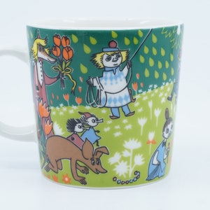 May include: White ceramic mug featuring a whimsical illustration with characters in a green field with flowers. The design includes a character holding a bouquet of orange flowers, and other figures with a dog. The mug has a white handle.