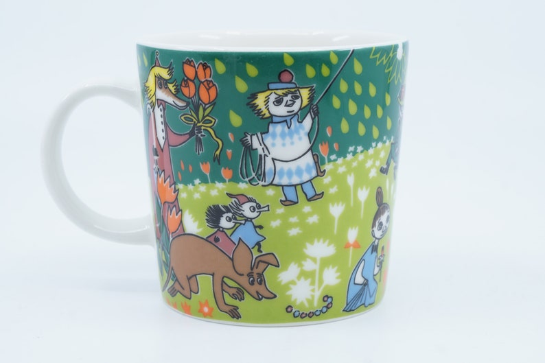 May include: White ceramic mug featuring a whimsical illustration with characters in a green field with flowers. The design includes a character holding a bouquet of orange flowers, and other figures with a dog. The mug has a white handle.