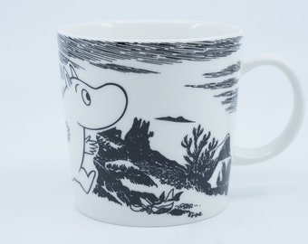 May include: White ceramic mug with a black and white illustration of a Moomin character looking up at a landscape. The illustration includes a mountain, trees, and a sky with clouds.