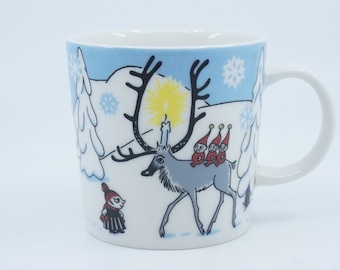 May include: White ceramic mug featuring a winter scene with a reindeer carrying three small figures. The reindeer has a candle on its antlers. Snowflakes and trees are in the background. A character stands on the left.