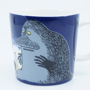 Può includere: Una tazza in ceramica blu con manico bianco presenta un'illustrazione di un cartone animato di una creatura blu con un naso grande e occhi gialli. La creatura tiene un oggetto bianco nelle sue mani. La tazza ha un bordo bianco e un interno blu.