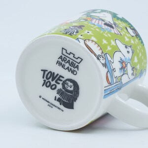 May include: White ceramic mug with a green and white illustrated design featuring characters. The bottom of the mug has the text "ARABIA FINLAND", "TOVE 100", and "MOOMIN CHARACTERS TM 2014". The mug has a white handle.