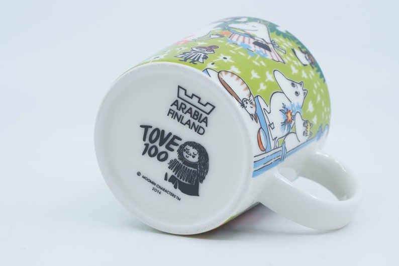 May include: White ceramic mug with a green and white illustrated design featuring characters. The bottom of the mug has the text "ARABIA FINLAND", "TOVE 100", and "MOOMIN CHARACTERS TM 2014". The mug has a white handle.