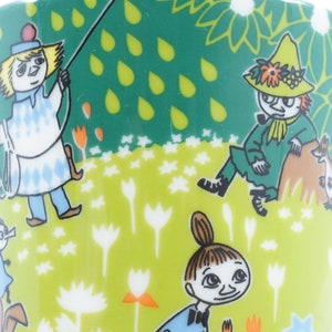 May include: A ceramic mug with a whimsical design featuring cartoon characters in a meadow setting. The mug has a white handle and a green background with floral and raindrop patterns. The characters are in various outfits.