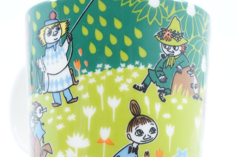 May include: A ceramic mug with a whimsical design featuring cartoon characters in a meadow setting. The mug has a white handle and a green background with floral and raindrop patterns. The characters are in various outfits.