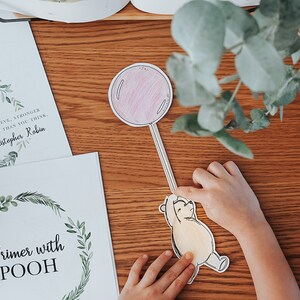 以下が含まれることがあります： 子供の手が、ピンク色の風船に手を伸ばすプーさんの切り抜きを持っています。緑の葉のリースとテキストが入った2つのプリントが木製の表面にあります。1つのプリントには「あなたはあなたが信じているよりも勇敢です...」と書かれており、もう1つには「Primer with POOH」と書かれています。
