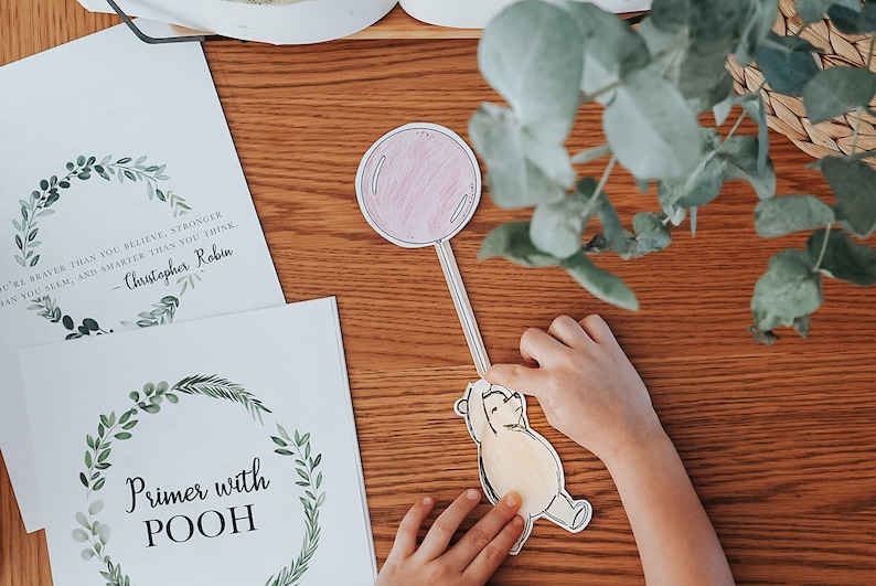 以下が含まれることがあります： 子供の手が、ピンク色の風船に手を伸ばすプーさんの切り抜きを持っています。緑の葉のリースとテキストが入った2つのプリントが木製の表面にあります。1つのプリントには「あなたはあなたが信じているよりも勇敢です...」と書かれており、もう1つには「Primer with POOH」と書かれています。