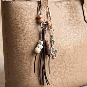 May include: A beige pebbled leather tote bag featuring a silver-toned metal keyring attachment. The keyring is adorned with two small wooden beads, a dark brown leather tassel, three white stone beads, and a silver-toned treble clef charm.