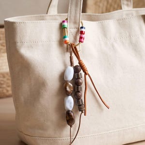 Peut inclure: Un sac fourre-tout en toile couleur crème doté d'un anneau en métal doré fixé à la poignée. L'anneau est orné de perles rayées colorées et d'une breloque composée de perles marbrées blanches et brunes et de perles texturées brun foncé.