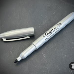 以下が含まれることがあります： キャップが外されたシルバーのSharpie Fine Point永久マーカーで、黒いペン先が見えています。マーカーは暗い表面にあります。「Sharpie」の文字がマーカーに印刷されています。