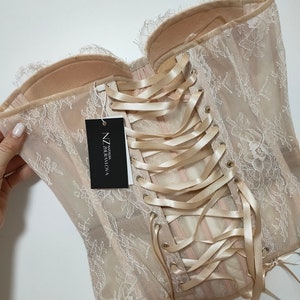 May include: A beige lace corset with a satin ribbon lacing up the back. The corset has a tag that reads "NZ Talia Graviova".