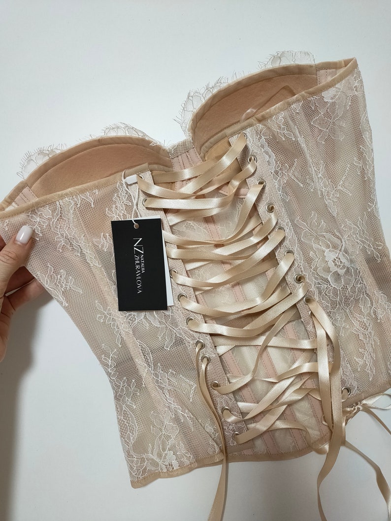 May include: A beige lace corset with a satin ribbon lacing up the back. The corset has a tag that reads "NZ Talia Graviova".