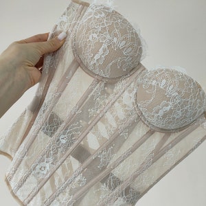 May include: A white lace corset with a beige mesh base. The corset has a sweetheart neckline and boning along the front.