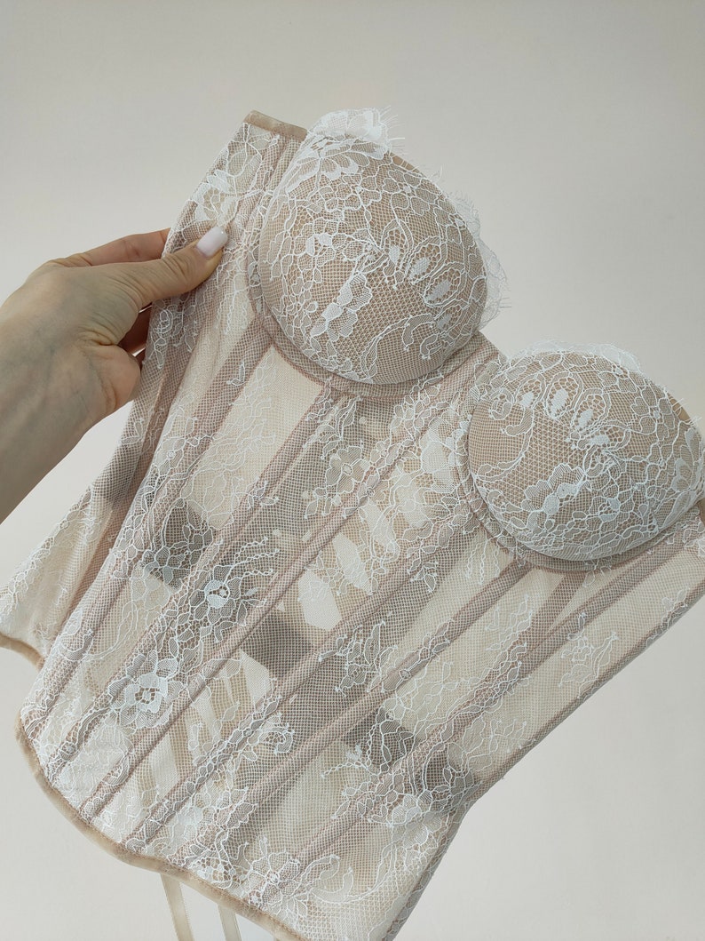 May include: A white lace corset with a beige mesh base. The corset has a sweetheart neckline and boning along the front.