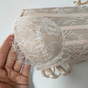 May include: A beige lace corset with white lace trim. The corset has a delicate floral pattern and is tied with a satin ribbon.