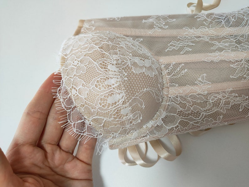 May include: A beige lace corset with white lace trim. The corset has a delicate floral pattern and is tied with a satin ribbon.