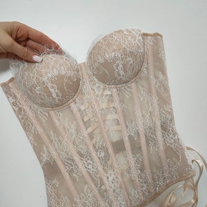 May include: A beige lace corset with white floral embroidery and satin ribbon ties. The corset has a boned structure and a sweetheart neckline.