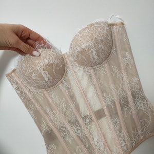 May include: A beige lace corset with white floral embroidery. The corset has a strapless design and a boned structure.