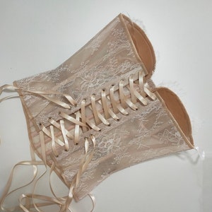 May include: A beige lace corset with a satin ribbon lacing system. The corset has a delicate floral pattern and is designed to be worn as a fashion accessory.