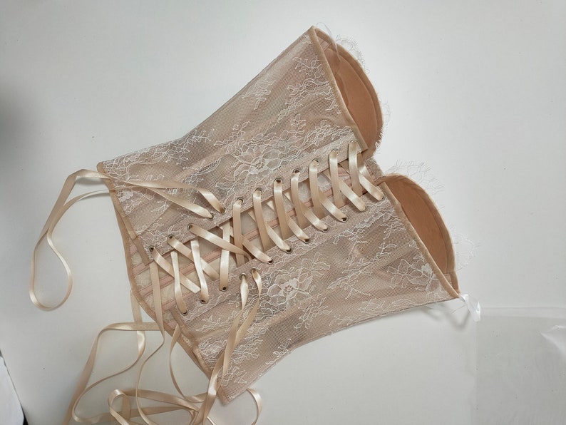May include: A beige lace corset with a satin ribbon lacing system. The corset has a delicate floral pattern and is designed to be worn as a fashion accessory.
