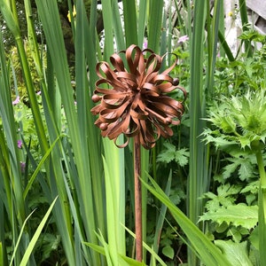 May include: A rusty metal garden sculpture in the shape of a flower with a long stem. The sculpture is surrounded by green grass and plants.