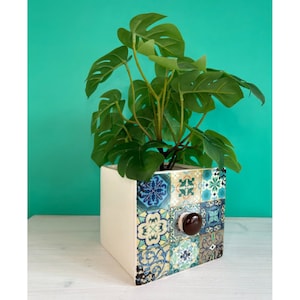 May include: A white ceramic planter with a blue, green and yellow geometric pattern. The planter has a brown knob and is filled with a green leafy plant.