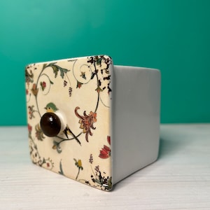 May include: A small white ceramic box with a floral design and a brown knob. The box is sitting on a white surface.
