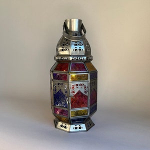 May include: A silver metal lantern with a colourful glass design. The lantern has a handle on the top and a geometric shape. The glass panels are in shades of red, yellow, blue, and purple.