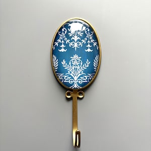 May include: An oval wall hook with a brass finish. The hook features a blue background with a white floral pattern. The hook is approximately 13 cm long.