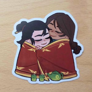 May include: A die-cut sticker featuring two cartoon characters with dark hair wrapped together in a large red blanket with gold trim. In front of them sit two small green tea cups and a matching green teapot with a circular design on the side.